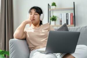 Asian woman wearing over-ear headphones feel bored while studying or working on a sofa in the living room at home. photo