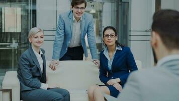 Team of cheerful business colleagues talking and discussing each other while having break sitting on sofa in modern office hall indoors photo