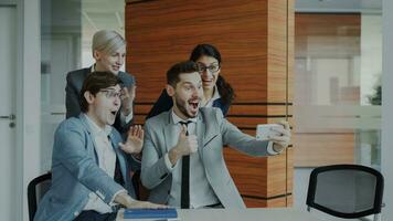 Happy business team taking selfie portrait on smartphone camera and posing for group photo during meeting in modern office indoors