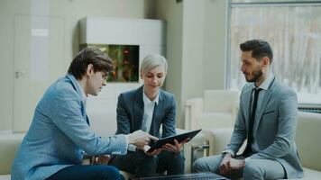 Panning shot of businessman discussing financial reports with female and male business partners sitting on sofa in modern office hall with indoors photo