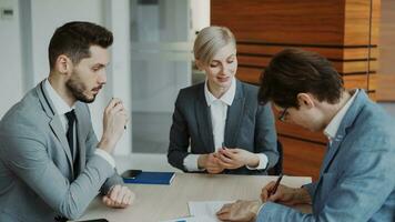 Young businessman in suit signing contract with his colleague while corporate lawyer explaining details of partnership sitting in modern office indoors photo