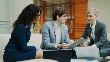 Businessman in glasses talking and duscussing future contract with female business partners sitting on couch in meeting room indoors photo