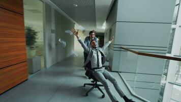 Two crazy businessmen riding office chair and throwing papers up while having fun in lobby of modern business center indoors photo