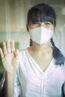 asiático adolescente vistiendo proteccion cara máscara en pie en hogar vivo habitación foto