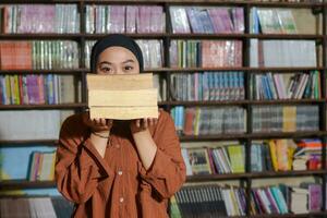 Portrait of Asian hijab woman covering her head with a pile of books in front of library bookshelf. Muslim girl reading a book. Concept of literacy and knowledge photo