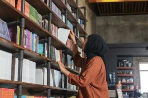 Portrait of Asian hijab woman holding book in front of library bookshelf. Muslim girl reading a book. Concept of literacy and knowledge photo