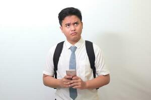 indonesio mayor alto colegio estudiante vistiendo blanco camisa uniforme con gris Corbata demostración Confusión cara expresión mientras participación un móvil teléfono. aislado imagen en blanco antecedentes foto
