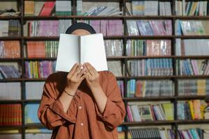 retrato de asiático hijab mujer cubierta su medio cabeza con un libro en frente de biblioteca estante para libros. musulmán niña leyendo un libro. concepto de literatura y conocimiento foto