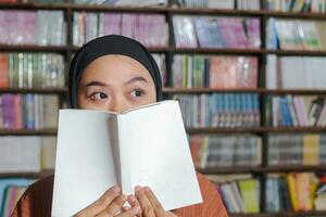 Portrait of Asian hijab woman covering her half head with a book in front of library bookshelf. Muslim girl reading a book. Concept of literacy and knowledge photo