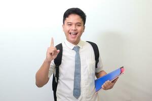 Indonesian senior high school student wearing white shirt uniform with gray tie holding some books, smiling and pointing up with finger reminding something. Isolated image on white background photo