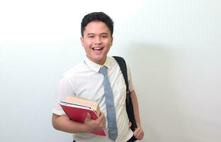 indonesio mayor alto colegio estudiante vistiendo blanco camisa uniforme con gris Corbata participación algunos libros, sonriente y mirando a cámara. aislado imagen en blanco antecedentes foto