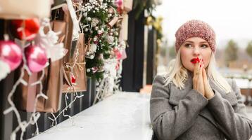 Gorgeous fashionable young blonde woman sitting near the window of the store or coffeehouse, dreaming, praying for a miracle before Christmas. cold winter day. photo