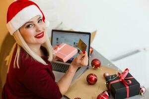 Smiling woman doing online Christmas shopping as she sits in front of the Xmas tree in a red Santa hat checking her card details to enter them onto her laptop computer. photo