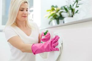 Woman in the kitchen is smiling and wiping dust using a spray and a duster while cleaning her house, close-up photo