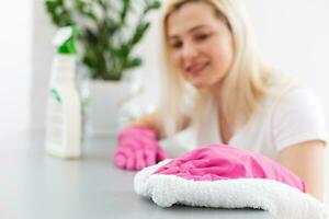 mujer en el cocina es sonriente y limpiando polvo utilizando un rociar y un plumero mientras limpieza su casa, de cerca foto
