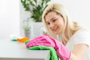 Woman in protective gloves is smiling and wiping dust using a spray and a duster while cleaning her house, close-up photo