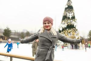 hermosa contento sonriente mujer a ciudad hielo pista en frente de Navidad árbol. contento joven adulto mujer disfrutando Navidad hora en el ciudad y mirando a cámara. foto