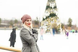 Beautiful happy smiling woman at city ice rink in front of Christmas tree. Happy young adult woman enjoying Christmas time in the city and looking at camera. photo