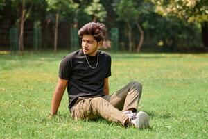 Attractive young indian man portrait in black t shirt and silver neck chain sitting on green lawn photo