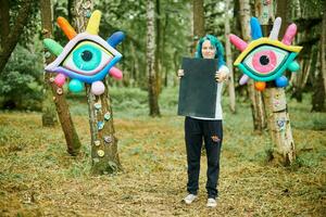 Funny young adult woman with dyed turquoise hair in white T shirt near big eye art installation photo