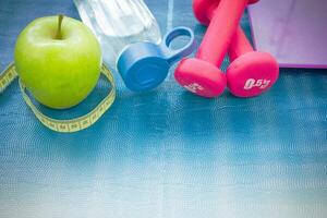 Two red dumbbells, an apple, a tape measure, a bottle of water and weight scales on a yoga mat photo