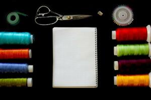 Multicolored threads, tape measure, scissors, pins and notepad for notes on a black background. View from above. photo