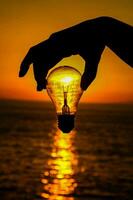 a person holding a light bulb over the ocean at sunset photo
