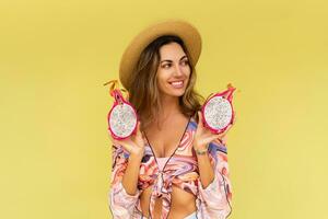Beautiful european woman in stylish summer outfit and straw hat posing on  yellow background. Fashion look. Holding tropical fruits. photo