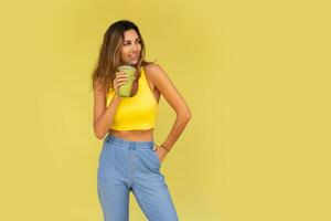 Studio photo of pretty brunette woman in sportive outfit  hoding beverage and posing on yellow background. Perfect slim body.