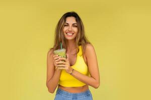 estudio foto de bonito morena mujer en juguetón atuendo hoding bebida y posando en amarillo antecedentes. Perfecto Delgado cuerpo.