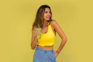 estudio foto de bonito morena mujer en juguetón atuendo hoding bebida y posando en amarillo antecedentes. Perfecto Delgado cuerpo.