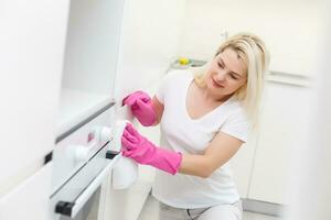 mujer en el cocina es sonriente y limpiando polvo utilizando un rociar y un plumero mientras limpieza su casa, de cerca foto