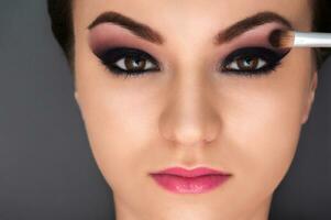 Close up shot of the young beautiful model with professional make up and pink lipstick and brush near her eye on the grey background photo