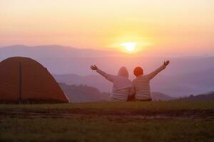 Pareja es sentado por el tienda durante durante la noche cámping mientras mirando a el hermosa escénico puesta de sol terminado el montaña para al aire libre aventuras vacaciones viaje foto