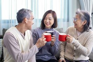 asiático familia de mayor padre madre y hija sentado en sofá con contento sonrisa en Jubilación hogar Bebiendo caliente té a celebrar su fiesta juntos para mayor cuidado y gasto valioso hora foto