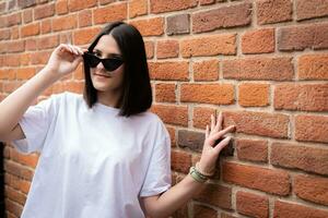 A cute brunette girl coquettishly adjusts her sunglasses against a brick wall. photo