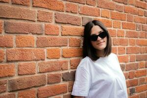 retrato de un morena en Gafas de sol soportes propensión espalda en contra un ladrillo pared foto