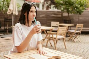 A young girl sits at a table in a summer cafe, drinks a drink through a straw and reads a book. photo