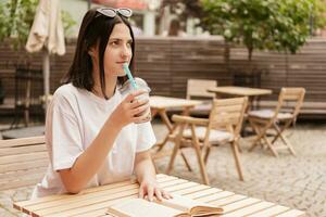 un joven niña se sienta a un mesa en un verano cafetería, bebidas un bebida mediante un Paja y lee un libro. foto