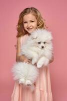 Little girl with a blond curly hair, in a pink dress plays with her dog photo