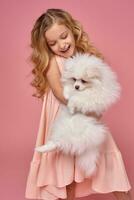 Little girl with a blond curly hair, in a pink dress plays with her dog photo