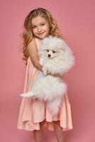 Little girl with a blond curly hair, in a pink dress plays with her dog photo