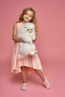 Little girl with a blond curly hair, in a pink dress plays with her dog photo