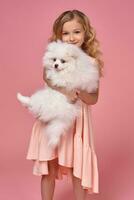 Little girl with a blond curly hair, in a pink dress plays with her dog photo