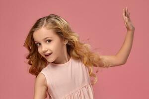 pequeño niña con un rubio Rizado cabello, en un rosado vestir es posando para el cámara foto