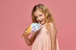 pequeño niña con un rubio Rizado cabello, en un rosado vestir es posando con un helado foto