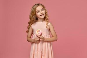 Little girl with a blond curly hair, in a pink dress is posing with a candy photo