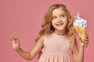 pequeño niña con un rubio Rizado cabello, en un rosado vestir es posando con un helado foto