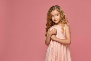 pequeño niña con un rubio Rizado cabello, en un rosado vestir es posando con un caramelo foto