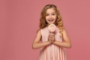 Little girl with a blond curly hair, in a pink dress is posing with a candy photo